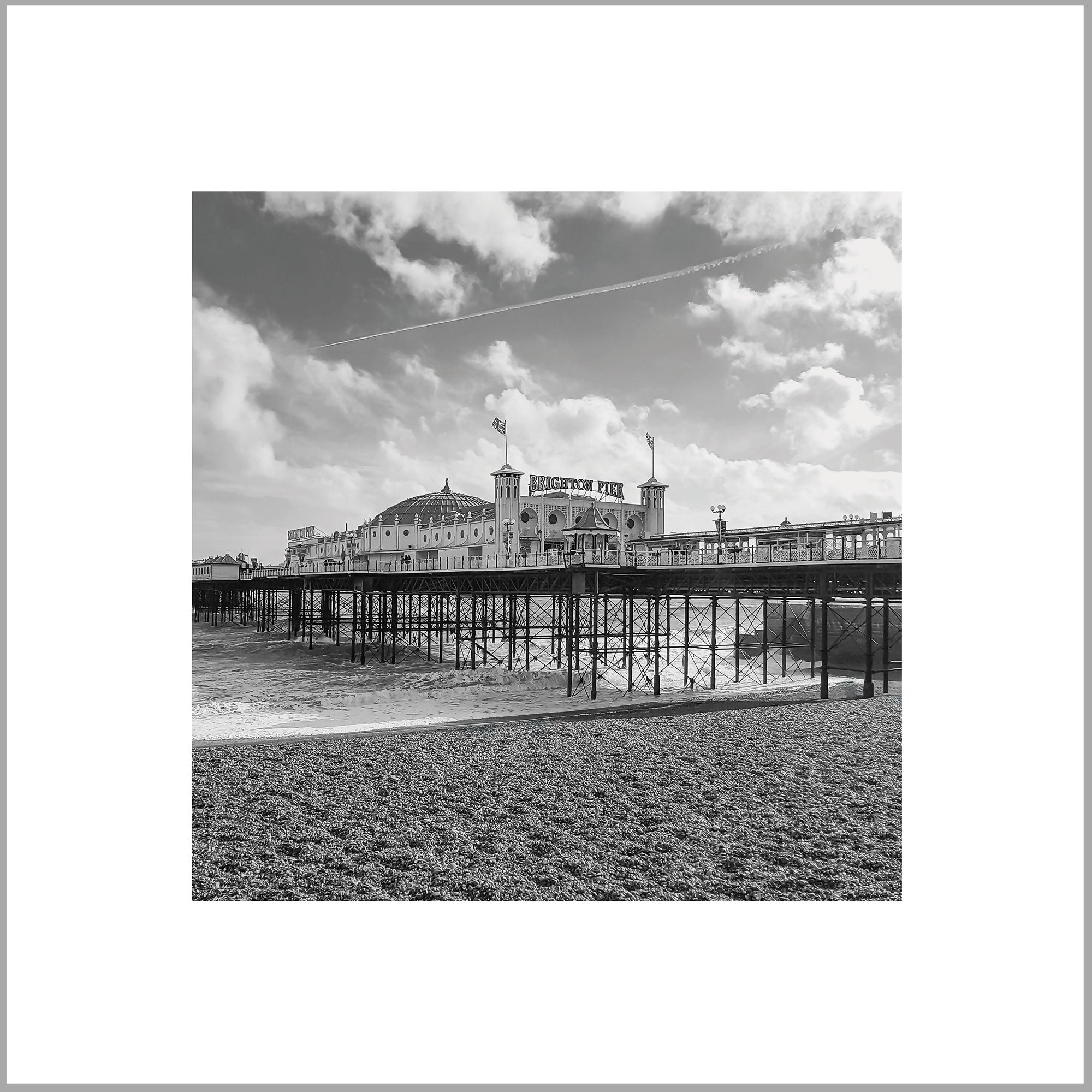 A black and white photograph of a pier extending into the water, with a large building or structure on top of it. The sky is cloudy, and there is a contrail visible in the distance.