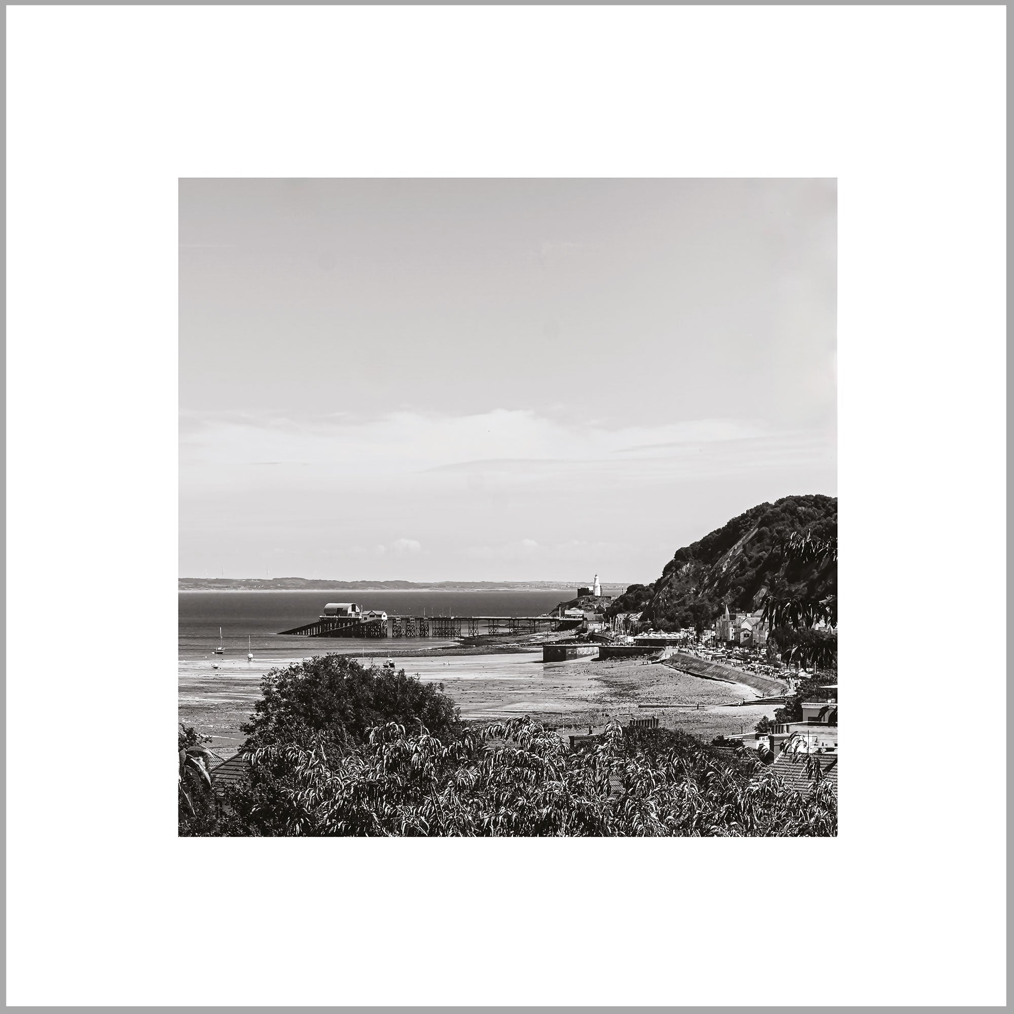 A black and white image of a coastal town with a pier and buildings along the shoreline, overlooking the ocean.