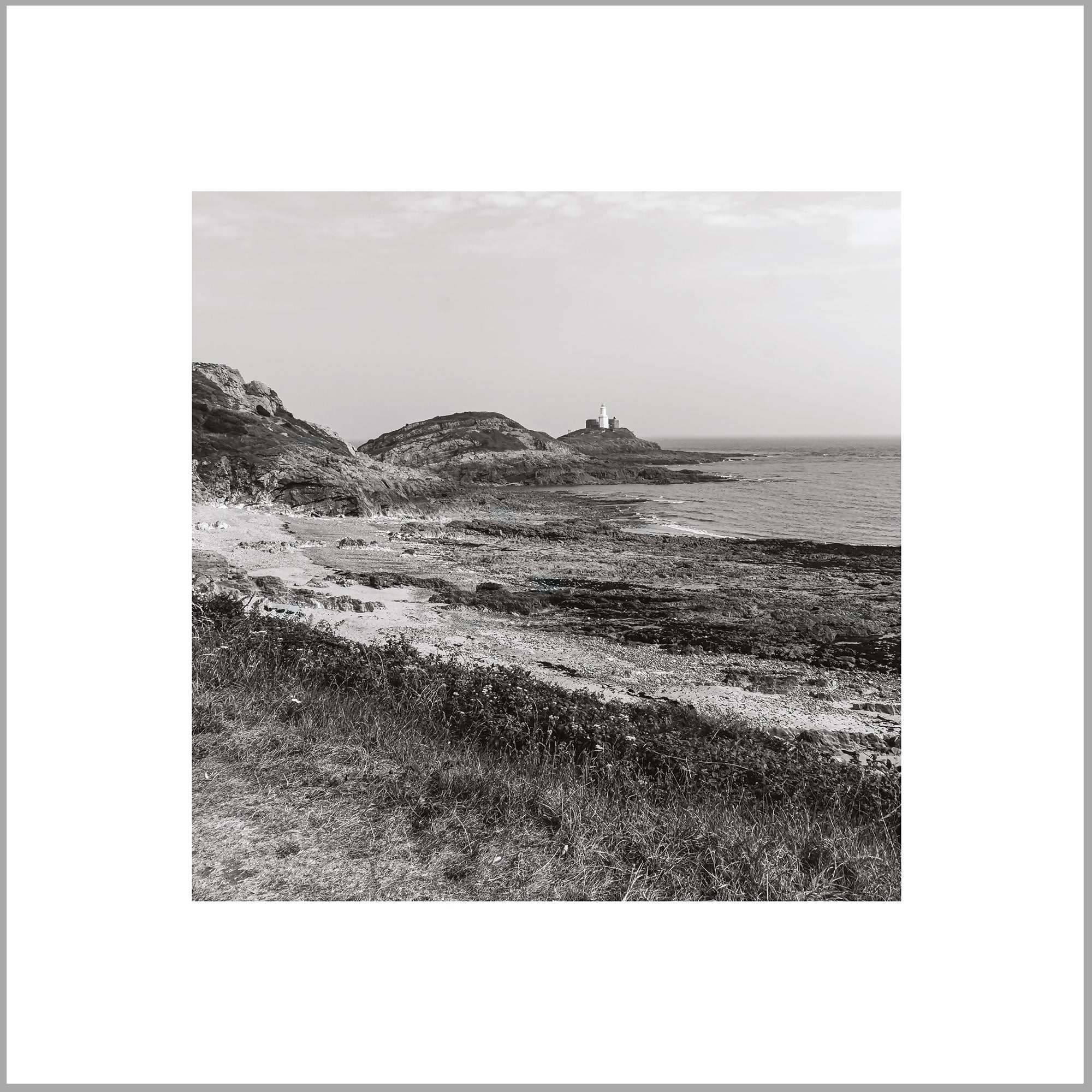 A black and white photograph depicting a rocky coastline with a lighthouse in the distance, overlooking the ocean.