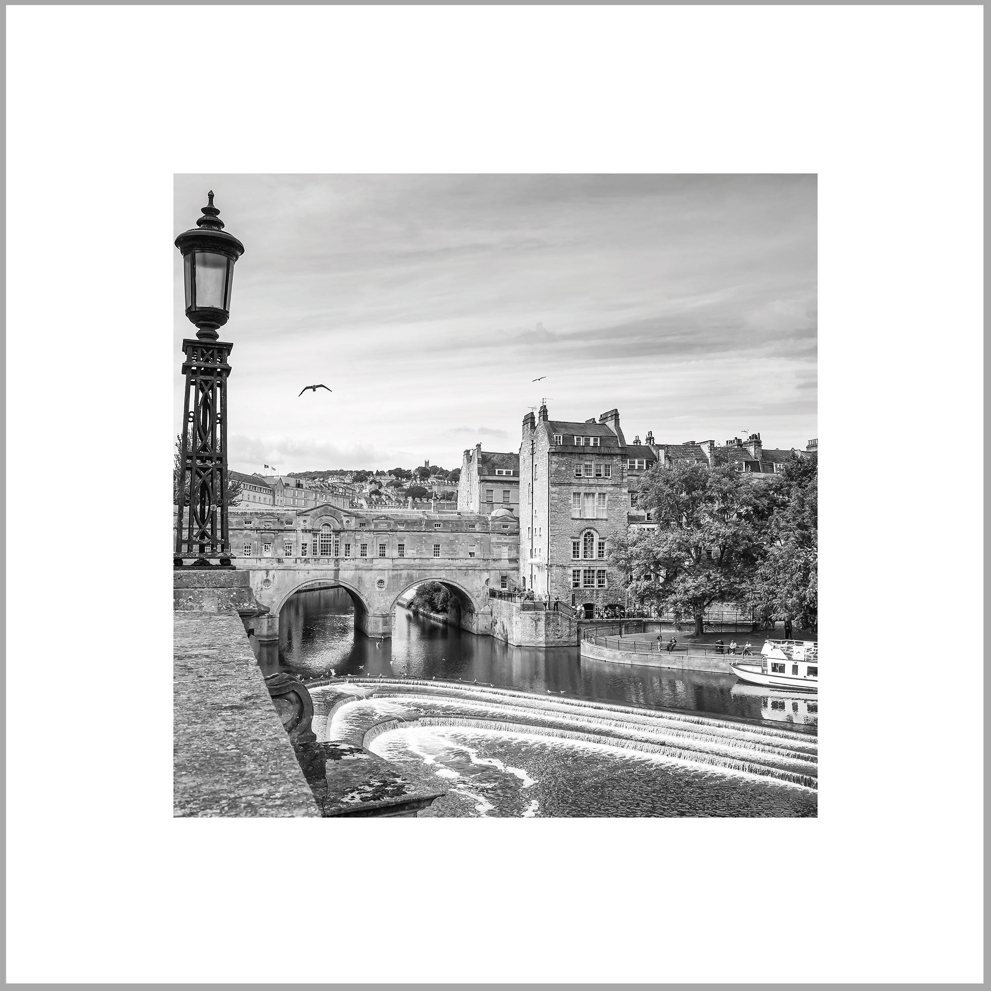 A black and white image of a cityscape with a bridge, buildings, and a river with a boat passing under it.