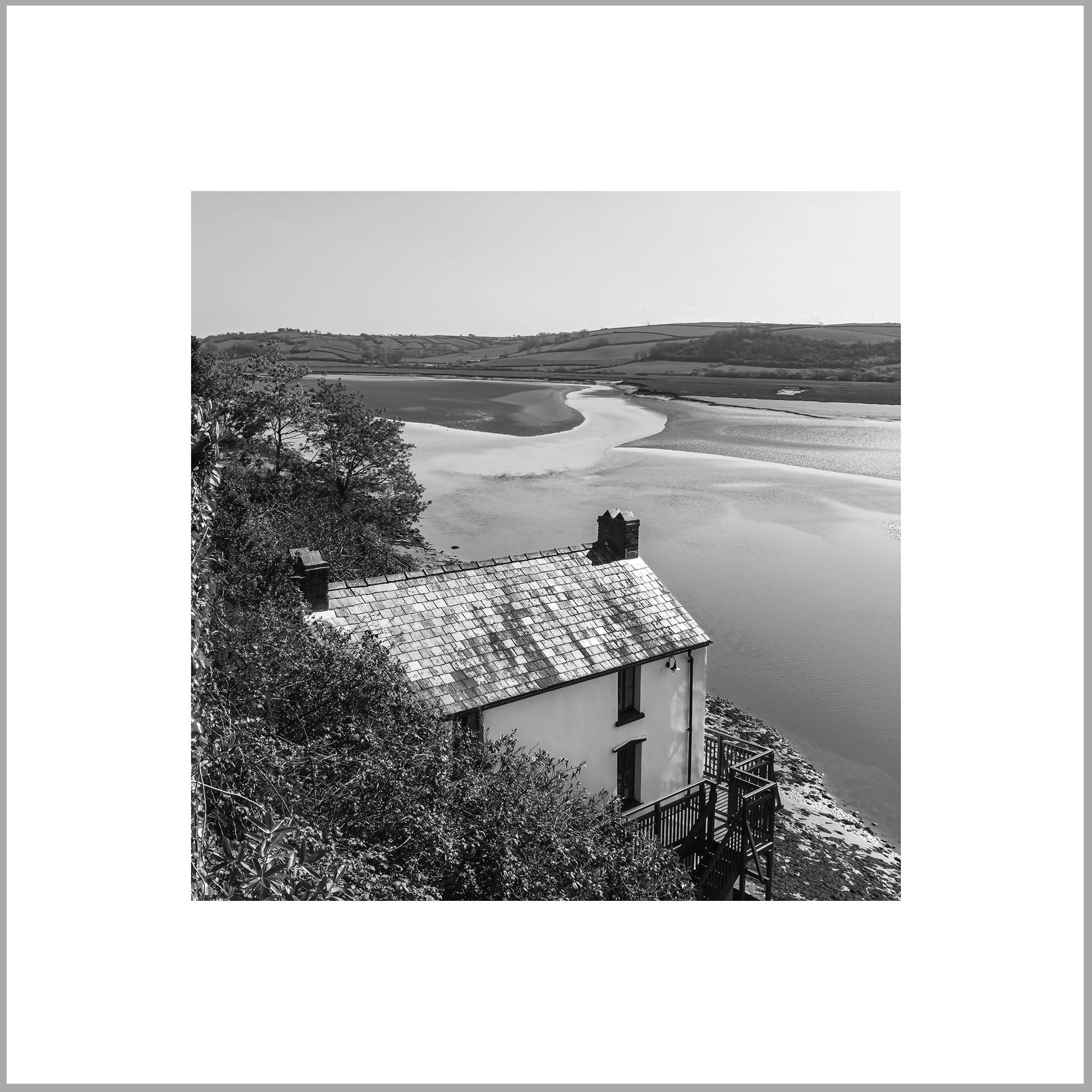A black and white image of a small house with a tiled roof, situated on a hill overlooking a body of water.