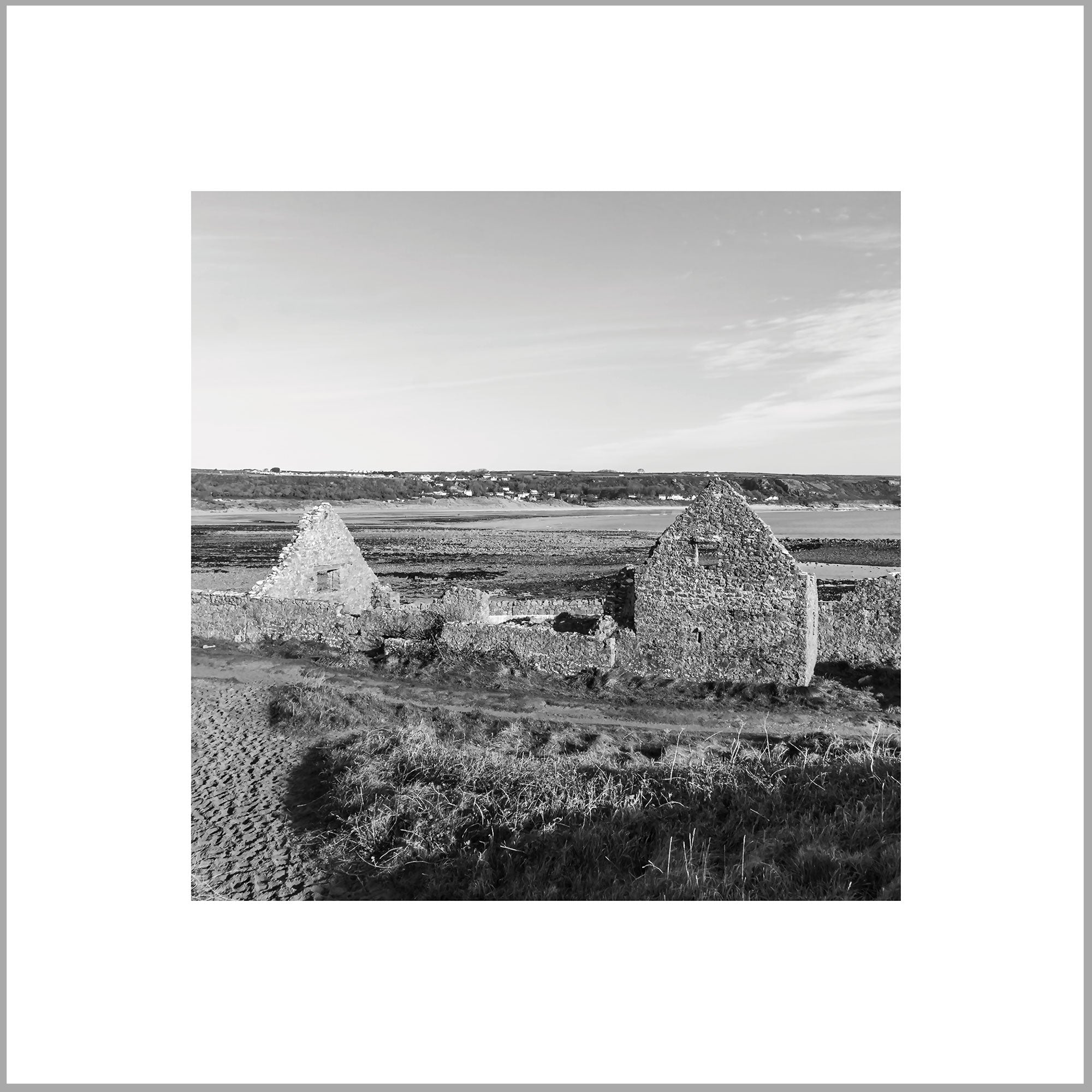 A black and white image showing two stone buildings in a rural setting, with a dirt road and grassy area in the foreground.