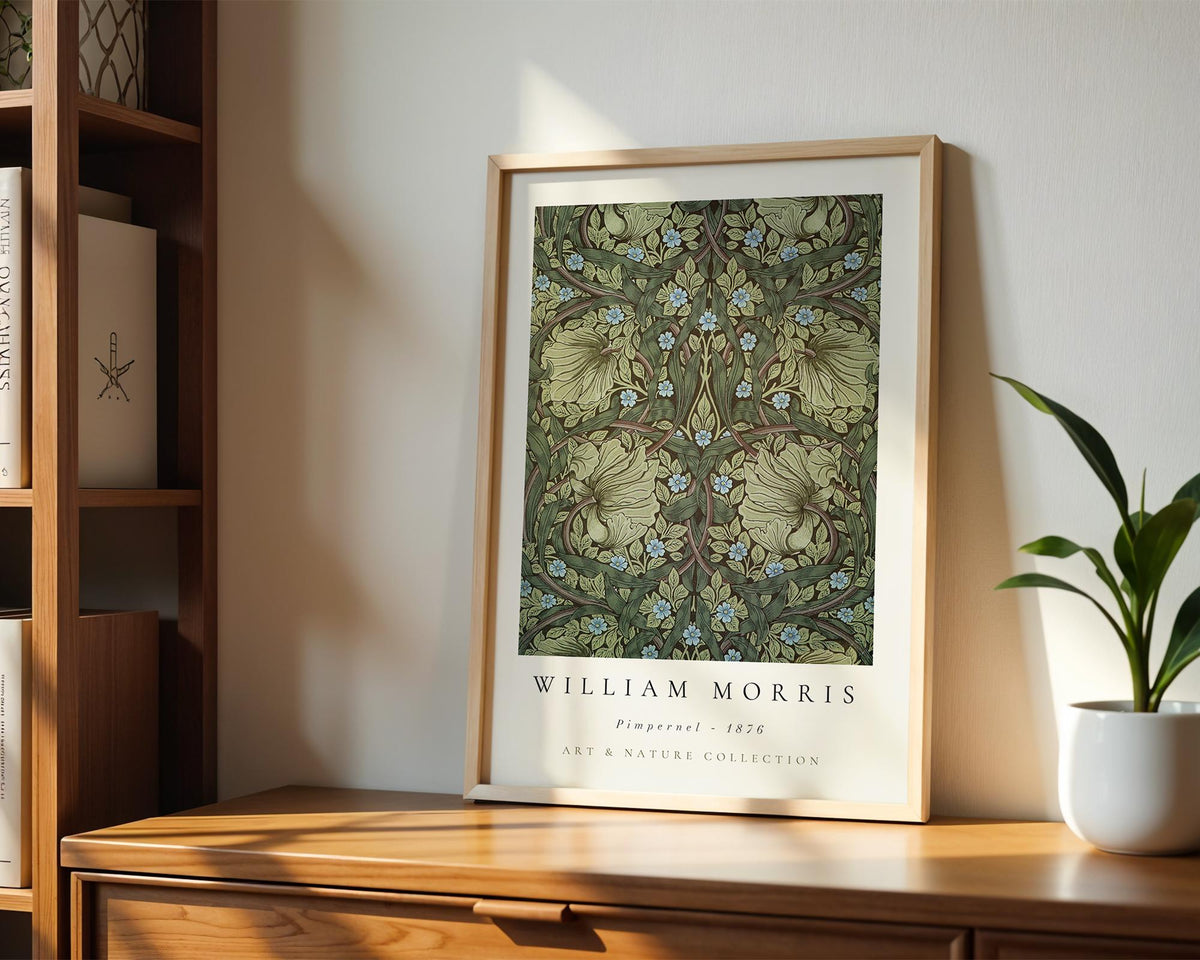 A framed print of William Morris' "Primerel 1856" art and nature collection is displayed on a wooden shelf, accompanied by a potted plant and a bookshelf in the background.