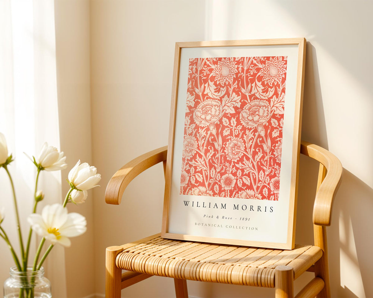 A framed print of William Morris' Pink & Rose pattern is displayed on a wooden chair, with a vase of white flowers to the left.