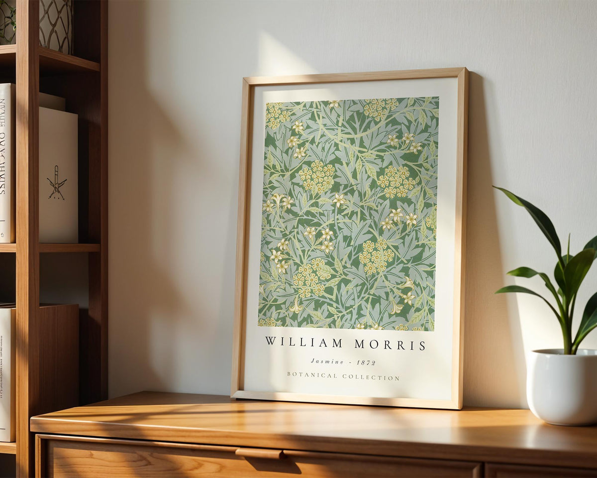 A framed poster of William Morris's "Jasmine - 1882" botanical collection is displayed on a wooden shelf, accompanied by a potted plant and a bookshelf in the background.
