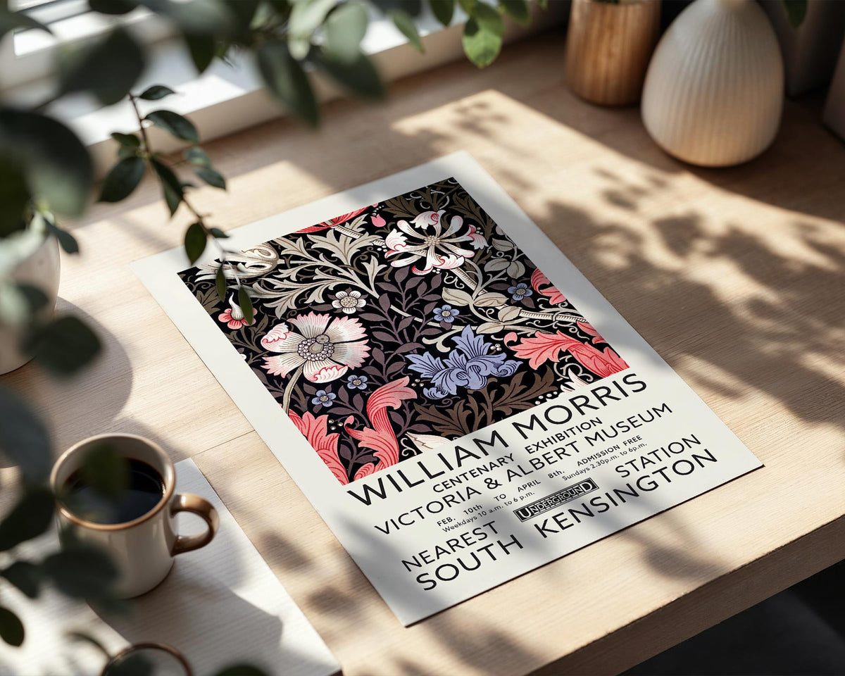 a poster advertising an exhibition of William Morris' work, placed on a wooden table next to a cup of coffee.